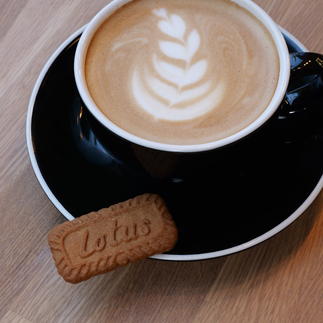 Eine Tasse Cappuccino mit Latte Art in Form eines Blattes, serviert in einer schwarzen Tasse mit passender Untertasse auf einem Holztisch. Daneben liegt ein Lotus Biscoff Keks.