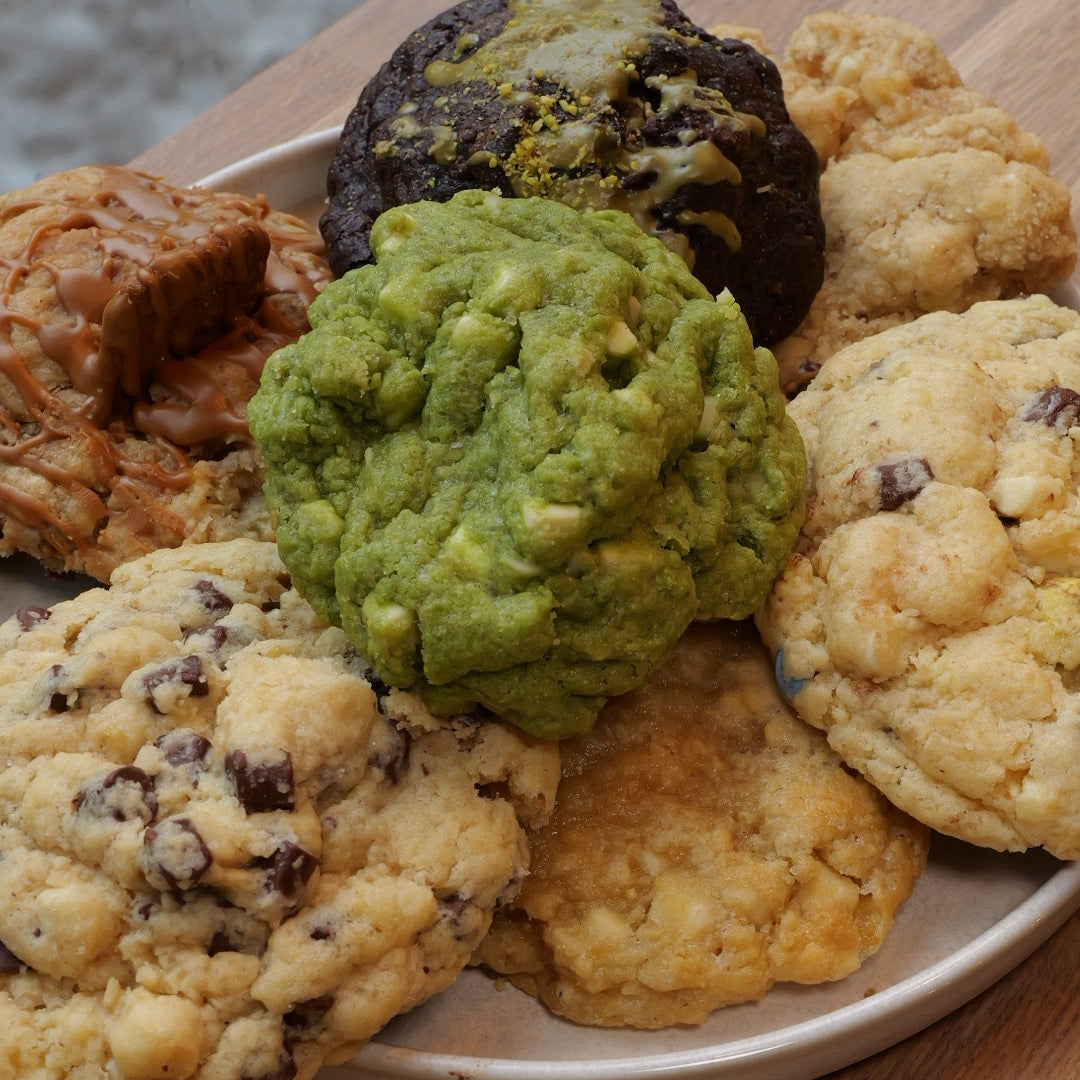 Ein Teller mit verschiedenen handgemachten Cookies auf einem Holztisch. Zu sehen sind Cookies mit Schokoladenstücken, ein grüner Matcha-Cookie mit weißer Schokolade, ein dunkler Schoko-Pistazien-Cookie, ein Lotus-Biscoff-Cookie mit Karamell und weitere Sorten mit unterschiedlichen Texturen und Toppings.