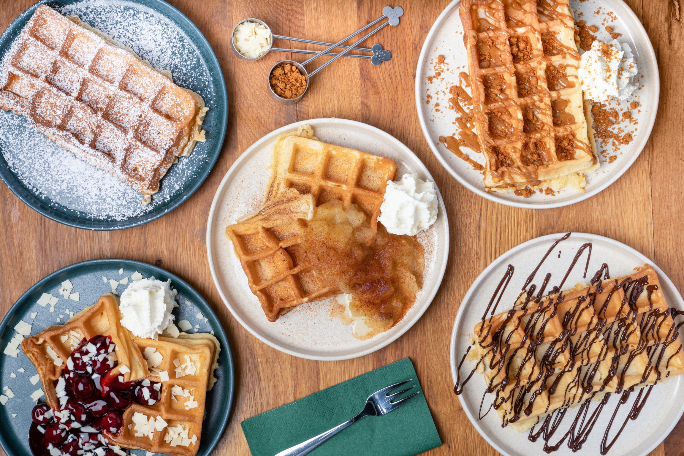 Fünf Teller mit frischen belgischen Waffeln, jeweils mit unterschiedlichen Toppings: Puderzucker, Kirschen und weißer Schokolade, Apfelmus und Zimt, Karamell mit Lotus Biscoff-Krümeln und Schokoladensauce. Dazu Schlagsahne als Beilage. Die Waffeln sind auf einem Holztisch angerichtet, mit kleinen Löffeln, die braunen Zucker und weiße Schokoladenraspeln enthalten.
