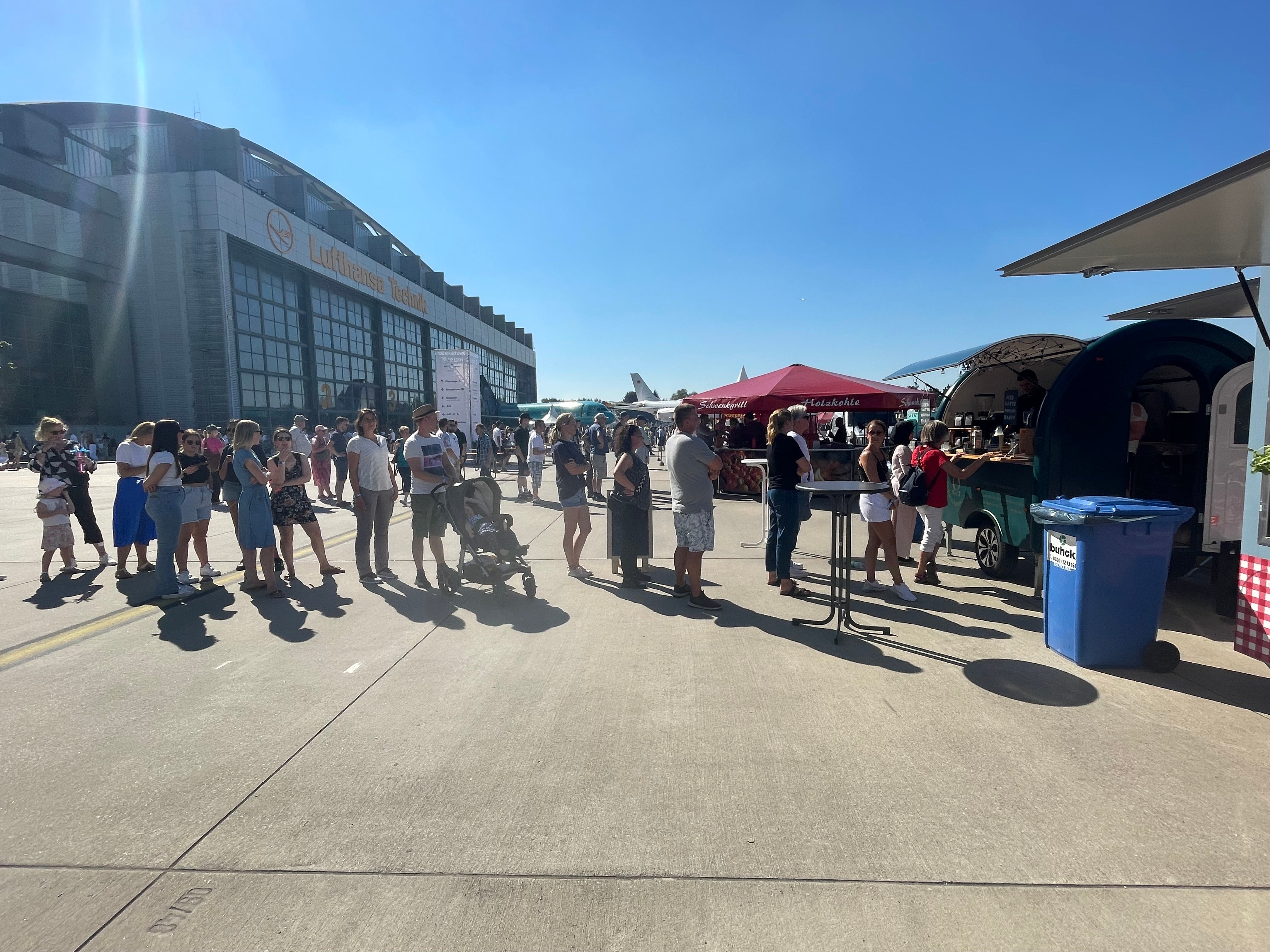 Kunden stehen in einer Schlange vor einem mobilen Kaffee-Catering-Wagen von Golden Beans unter freiem Himmel bei einem Event vor der Lufthansa Technik Halle.